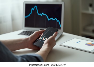Young faceless man analysing graphs and data on computer and smartphone at office. Online trader and stockholder, gen Z businessman using app on mobile and buying stocks. Trader or investor concept