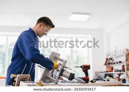 Young factory worker assembling machine part in bright workshop, wearing protective glasses and gloves, working on metal object