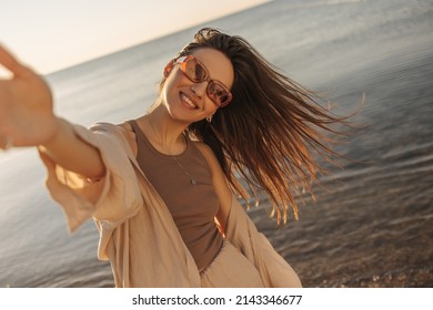 Young fair-skinned happy lady makes selfie in sunglasses on background of sea. Brown-haired woman is waving her hair in upbeat cheerful mood. Leisure, relaxation, activity and lifestyle concept.