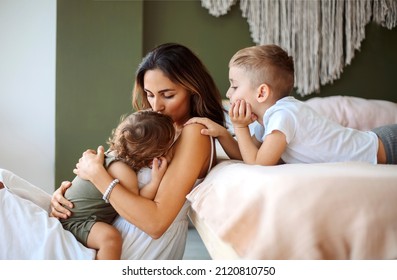 Young family beautiful mother with two son of different ages spending time at home, loving mom holding baby in hands and putting him to sleep with help and support of older child