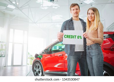 Young Family Buying First Electric Car In The Showroom. Smiling Attractive Couple Holding Paper With Word Credit While Standing Near Eco Red Vehicle. Battery Electric Car For Ecology.