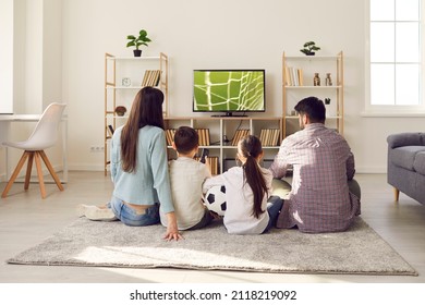 Young family with children watching football match together at home cheering for their favorite team. Mom, dad, daughter and son are sitting on floor in front of big TV with their backs to camera.