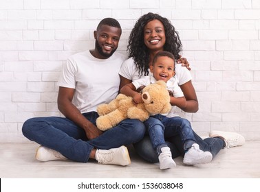 Young Family Concept. Happy Black Parents With Little Son Sitting On Floor Together, Smiling And Posing To Camera Over White Brick Wall Background, Free Space