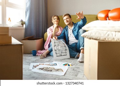 Young Family Couple Bought Or Rented Their First Small Apartment. People Sit Together On Floor. Guy Point Forward. He Hold Apartment Plan. Young Woman Smile.