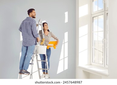 Young family couple doing renovations and painting walls inside house. Happy, joyful man and woman standing on ladder at home, holding bucket and rollers and looking at light walls and sunny windows - Powered by Shutterstock