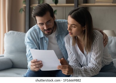 Young Family Couple Engaged In Reading Loan Contract Mortgage Agreement Look Through Terms Conditions Think On Signing Paper. Pensive Spouses Husband Wife Embrace On Sofa Study Legal Document Hardcopy