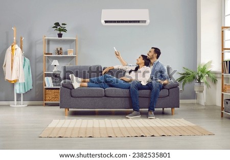 Young family couple enjoying life in their apartment with a modern air conditioning system. Happy husband and wife relaxing on the sofa at home and setting up the temperature on their AC on the wall