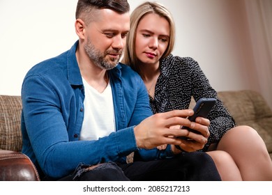 Young Family Couple Friends Sitting On Couch Hold Phones Discuss Apps, Sharing Information Between Gadget Together