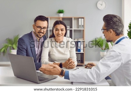 Young family couple planning children and visiting doctor. Doctor talking to happy husband and wife, explaining details of IVF treatment, pointing at laptop computer and showing something on screen