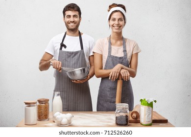 Young family couple standing on kitchen, cooking dinner together, surrounded with different products and tablewear, having happy expressions. Joyful man and woman preparing pancake for breakfast - Powered by Shutterstock