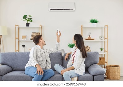 Pareja joven de la familia que enciende el aire acondicionado mientras está sentado en el sofá en la sala de estar. Hombre y mujer felices usando el control remoto, configurando la temperatura en el sistema de CA, disfrutando del aire fresco en casa.