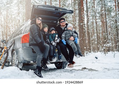 Young Family Dressed In Warm Clothing Pose On Camera Sitting On Car Trunk In Winter Forest In Daytime.