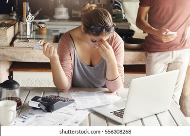 Young Family Facing Debt Problems, Can't Pay Out Their Mortgage. Stressed Woman Holding Her Head In Frustration, Doing Bank Accounts And Making Necessary Calculations Using Laptop And Calculator