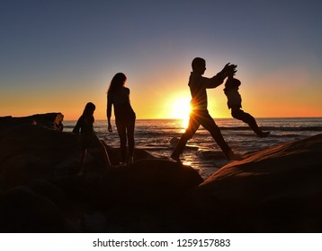 A Young Family Of Four Enjoying A Sunset Walk Along The Cliffs At A San Diego Beach. The Father Is Lifting His Young Son Across The Boulders.