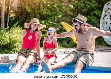 Young Family Having Fun By Swimming Pool.Happy Young Family Splashing Water With Legs While Sitting On Edge Of Swimming Pool.