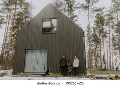 Young Family With Kids And Dog Standing In Front Of Purchased New Barn Style Black House. Modern Countryside Architecture. Affordable Real Estate With Bank Mortgage Loan. Family Escape From Virus.