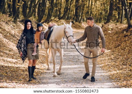 Young family is learning to ride a beautiful white horse in the autumn forest. Riding lesson