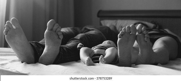 Young family lie on a bed feet to the viewer. The family consists of mother, father and little baby. Black-and-white background picture. 