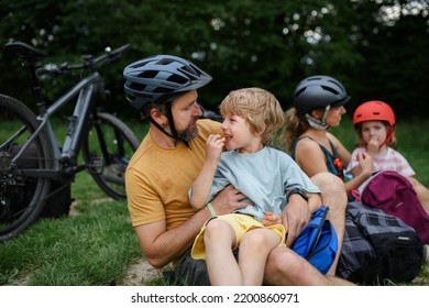 Young Family With Little Children Resting After Bike Ride, Sitting On Grass In Park In Summer.