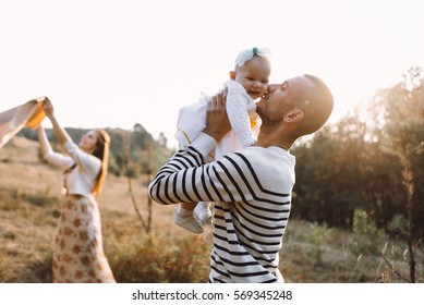 Young Family With Little Daughter Walking Along The Mountain Slopes On The High Yellow Grass. Dad Throws Daughter Into The Sky Backlit Sunlight 