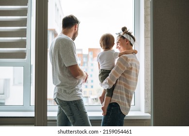 Young Family Man Father And Mother Woman With Baby Girl On Window Sill Looking At Window At Home