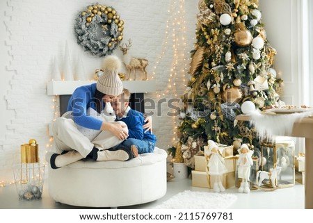 A young family - mom and son, sitting in a bright Christmas decorated room in gold tones and tenderly embracing, holding a present with a live white rabbit in their hands.