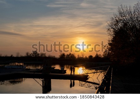 A young family with a mother, father, and small baby stand on a dock with their reflections in the water below, enjoying a beautiful sunset at Colonel Samuel Smith Park in Toronto (Etobicoke), Ontario