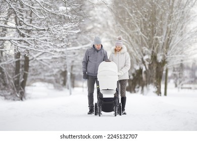 Young family pushing baby stroller and walking on snow covered sidewalk at park in cold winter day. Enjoying peaceful stroll. Spending time with newborn and breathing fresh air. Front view.