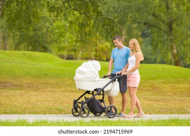 Young Family Pushing White Baby Stroller And Walking On Sidewalk At Beautiful Town Park. Warm Sunny Summer Day. Spending Time With Newborn And Breathing Fresh Air. Enjoying Peaceful Stroll. Side View.