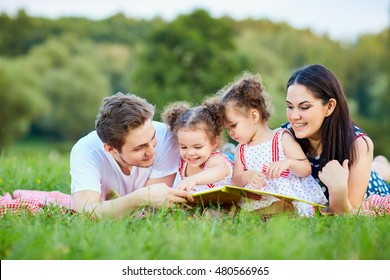 Young Family Reading A Book Outdoors Lying On The Grass In The Park.