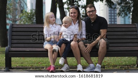 A young family is sitting on a park bench and having fun. Happy diti with parents relax outdoors in summer.
