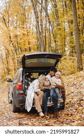 Young Family Sitting At Open Trunk Of Hatchback Car In Autumn Forest