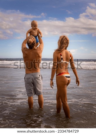 Young family spending summer vacation in Asia. Father holding, lifting his infant baby boy son high in the air. View from back. Family beach concept. Blue sky, white clouds. Seminyak beach, Bali