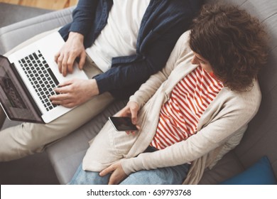 Young Family Spending Time With Electronic Devices. Man Using Computer And Smiling Woman Typing Message With Mobile Phone. Sitting Together On The Sofa