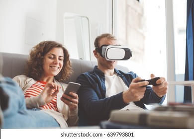 Young Family Spending Time Together With Electronic Devices. Man Playing Breathtaking Console Game With VR Headset. Happy Woman Typing Message Using Mobile Phone