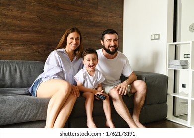 A Young Family Spends Home Leisure Watching TV. Happy Mom, Dad And Little Son Are Sitting Behind A Sofa In A Cozy Living Room. Photo With Grain