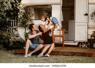 A Young Family Spends A Summer Evening Near Their Trailer. Mobile Home As A Way Of Comfortable Family Travel.Happy Young Mother Holds Little Daughter In Her Arms. Husband Hugs His Wife