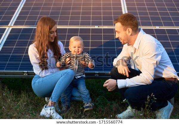 Young family of three is crouching near\
photovoltaic solar panel, little boy is looking at camera, parents\
looking at him, modern family\
concept