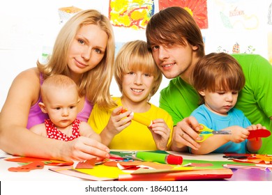 Young family with three kids sitting by the table and crafting cutting and gluing paper