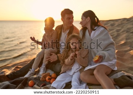 A young family with two children sitting on a blanket on a picnic on a sandy shore by the sea at sunset having fun and relaxing on the weekend.