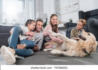 Young Family With Two Kids Sitting On Floor In Living Room And Playing With Dog