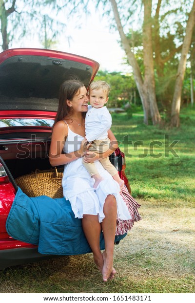 Young family in white clothes beautiful woman\
mother embracing her son in park in front of trunk car. Summer\
holidays. Happy scene
