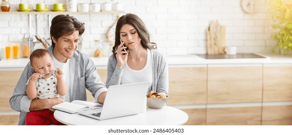 A young family is working from home in their kitchen. The mother is on the phone while the father is holding their baby and working on a laptop - Powered by Shutterstock