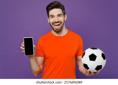 Young fan man he wear orange t-shirt cheer up support football sport team hold soccer ball watch tv live stream use mobile cell phone blank screen workspace area isolated on plain purple background. - Shutterstock ID 2176696389
