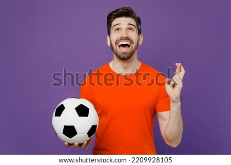 Young fan man he wears orange t-shirt cheer up support football sport team hold in hand soccer ball watch tv live stream wait special moment, keep fingers crossed isolated on plain purple background