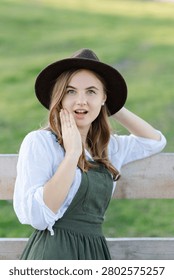 Young farmer with hat looking surprised touching her face and adjusting her hat standing next to a wooden fence in a field
