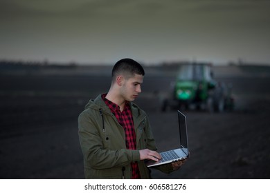 Young farmer with laptop standing in field with tractor harrowing in background