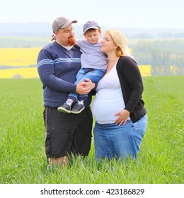 Young Farmer's Family Together On Green Wheat Field. Pregnant Mother And Overweight Father With Their Little Son. People Working And Enjoying Life On Countryside. Woman, Man And Child.