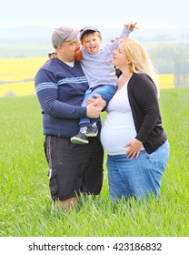 Young Farmer's Family Together On Green Wheat Field. Pregnant Mother And Overweight Father With Their Little Son. People Working And Enjoying Life On Countryside. Woman, Man And Child.