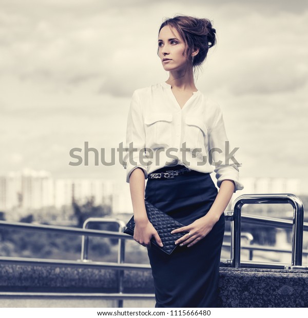Young fashion business woman with clutch bag walking on\
city street  Stylish female model in white shirt and dark blue\
pencil skirt 